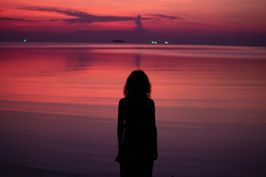 Silhouette of a woman standing by the sea at sunset with pink and orange hues reflecting on the water.